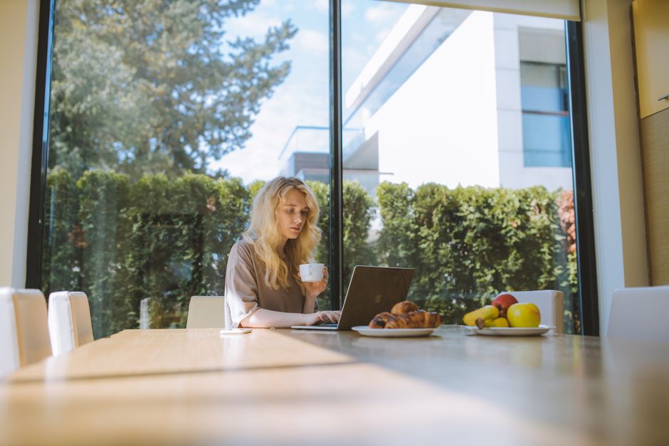 Blonde woman working on a laptop at home, enjoying a coffee with croissants